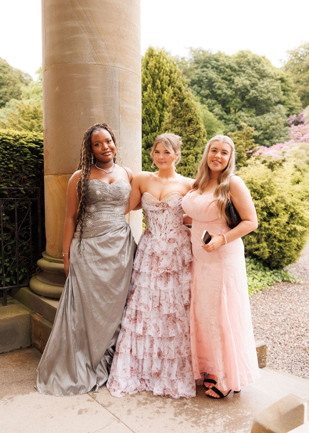 Three young women in elegant formal gowns pose together outdoors near a stone column at prom.