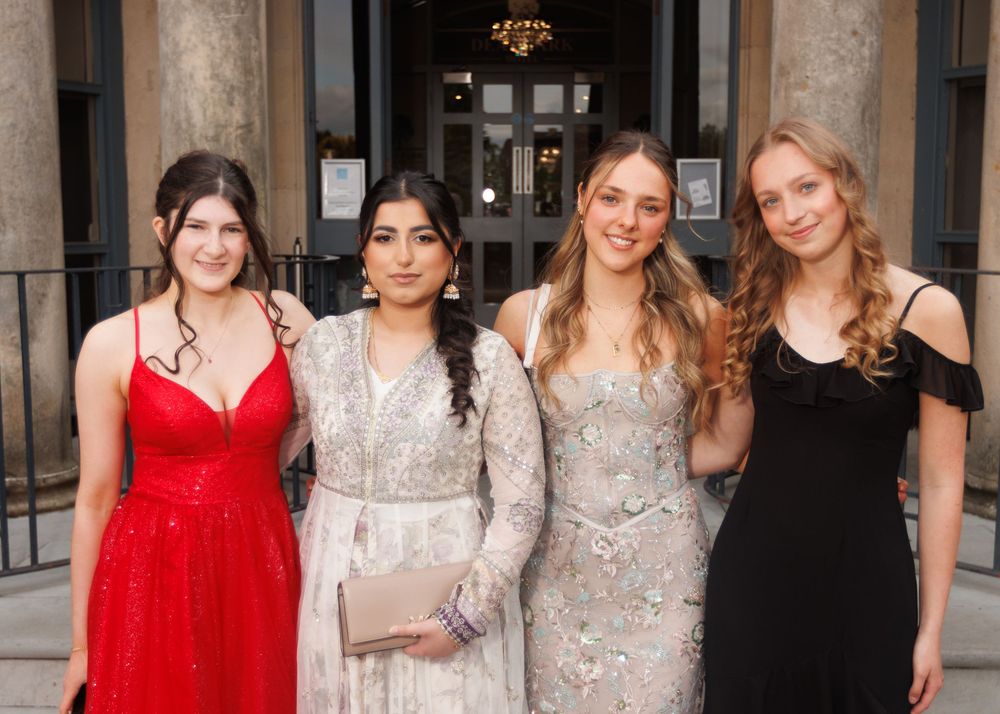 Four friends dressed up for a formal occasion pose together wearing elegant dresses in red, silver, sparkly beige and black.
