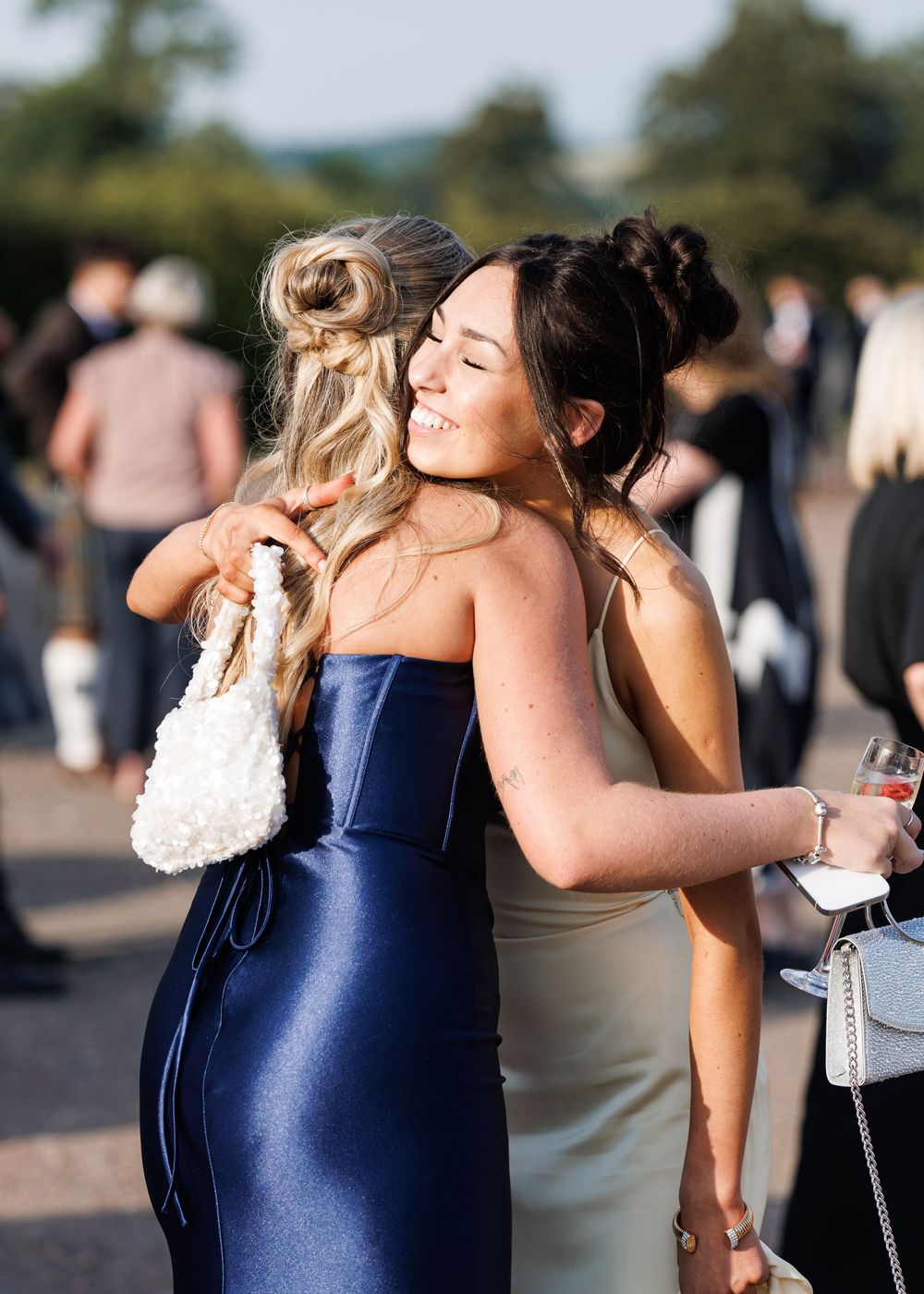 Two women in elegant satin dresses embracing warmly at an outdoor formal event.