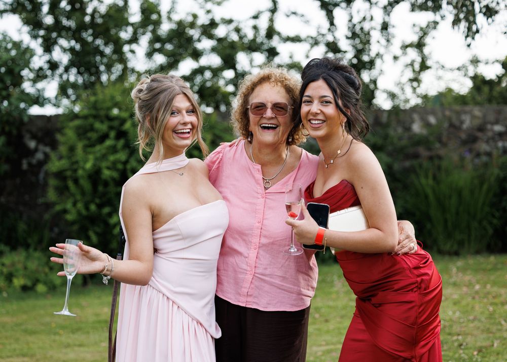 Three women smiling and posing together outdoors at a garden event, dressed in elegant pink and red formal attire.