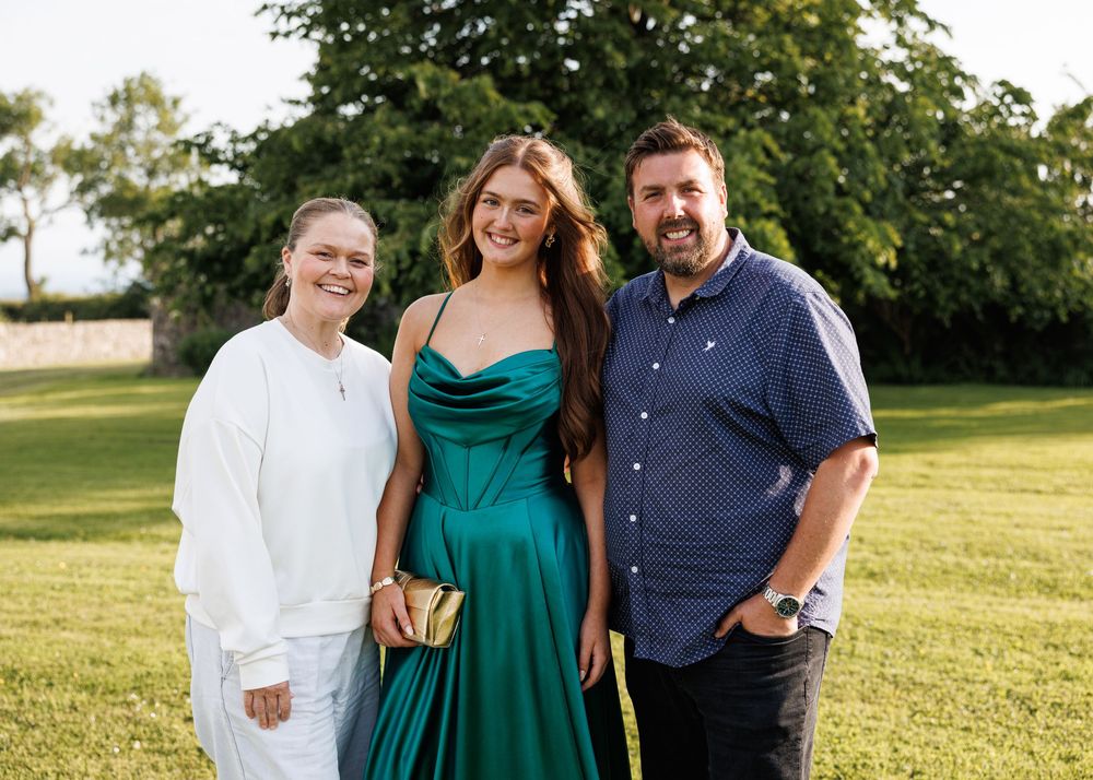 A young woman in a green satin gown poses with two adults on a grassy lawn for a formal occasion.