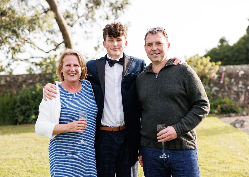 Family of three smiling outdoors at a formal event, holding champagne glasses in a lush garden setting.