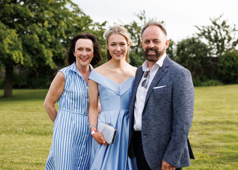 Three people smiling outdoors on green grass, dressed in elegant blue outfits for a formal occasion.