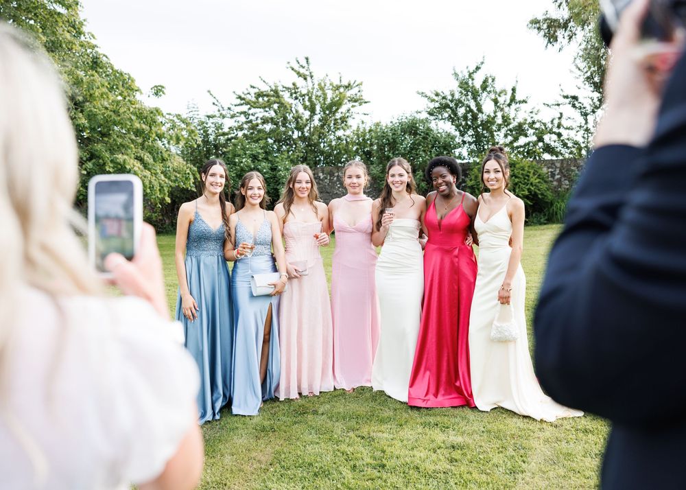 Six bridesmaids in colorful floor-length gowns pose outdoors while guests photograph them at a wedding.