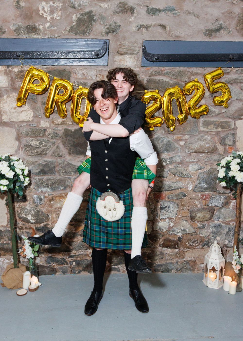 Person in traditional Scottish attire including a green tartan kilt and black vest poses playfully against a stone wall.