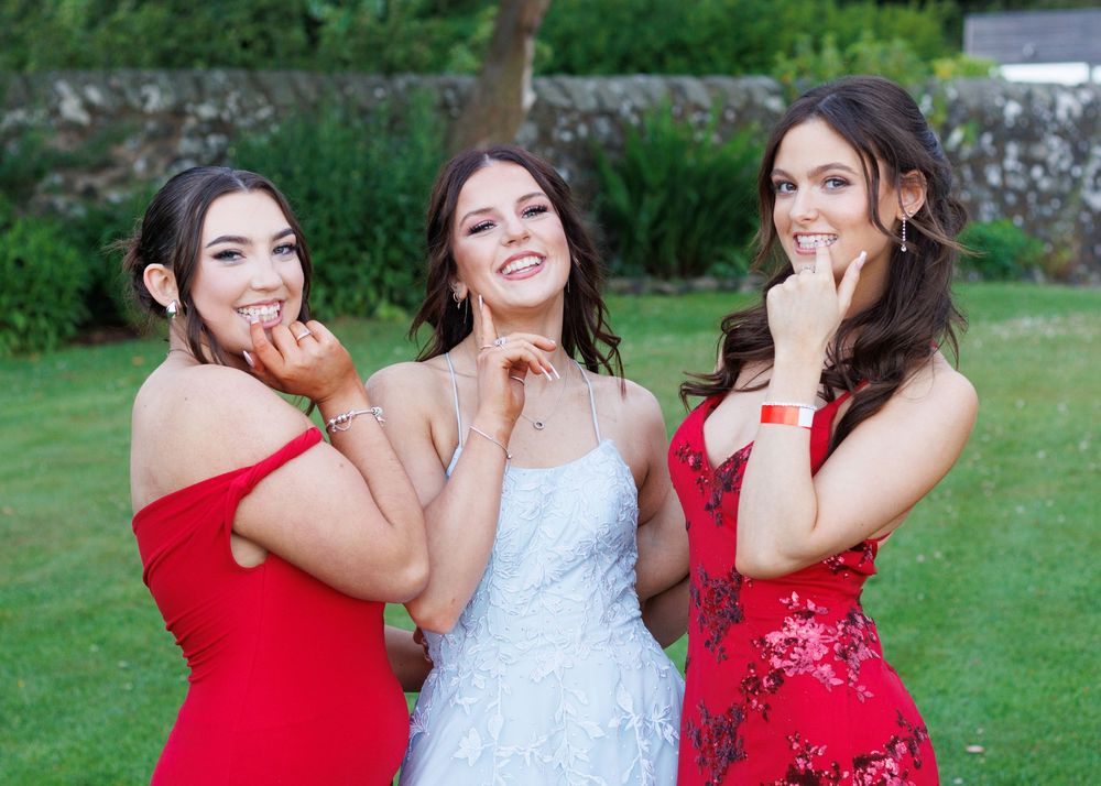 Three women in formal dresses pose together playfully on a green lawn at an outdoor celebration.