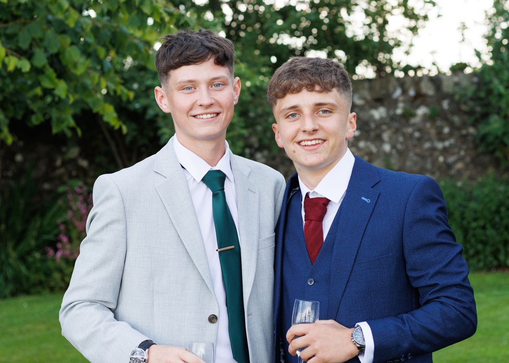 Two people in formal attire smile and pose together while holding wine glasses in an outdoor garden setting.