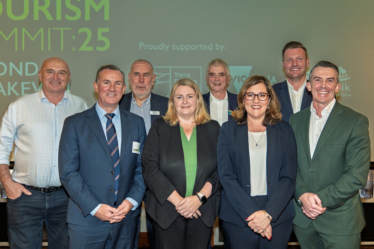 Group of nine business professionals pose together at Tourism Summit 25 conference in a modern venue.