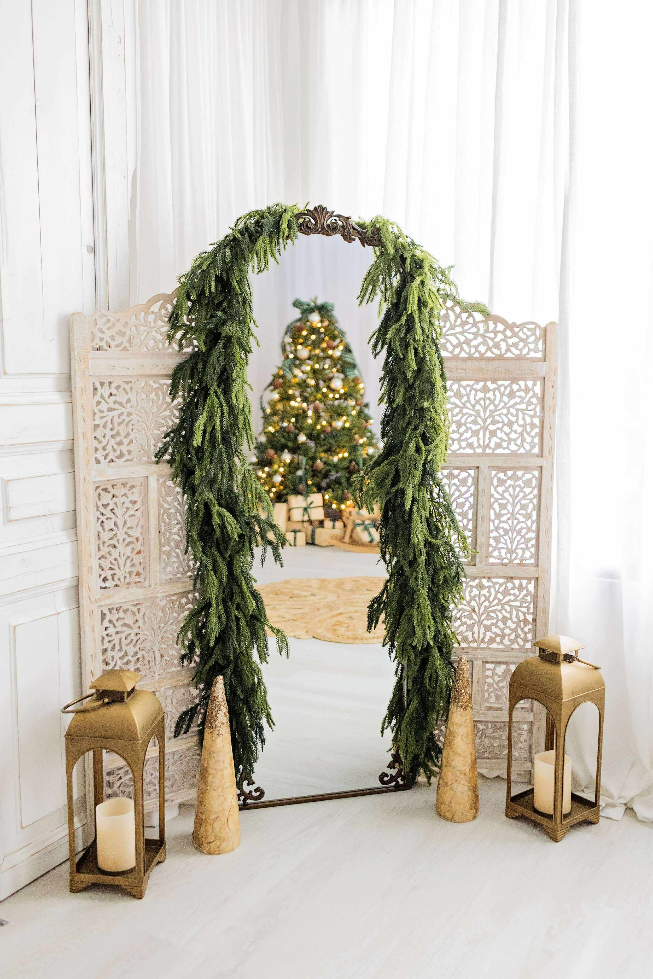 Elegant mirror framed with fresh greenery garland and flanked by gold lanterns reflects a lit Christmas tree.