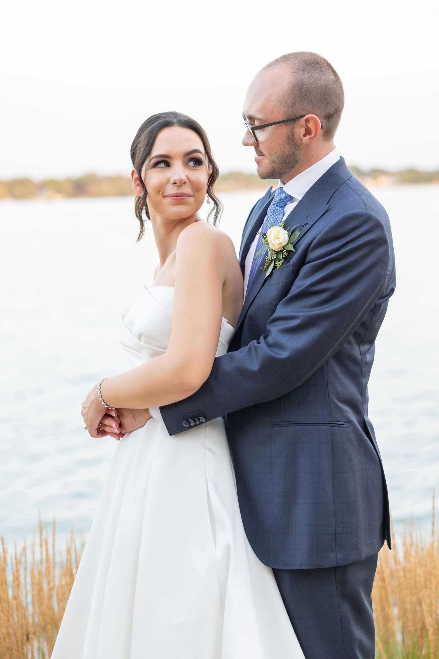 A romantic newlywed couple embraces by a waterfront with golden grass at sunset, the bride in white and groom in navy. Rachel Martinez