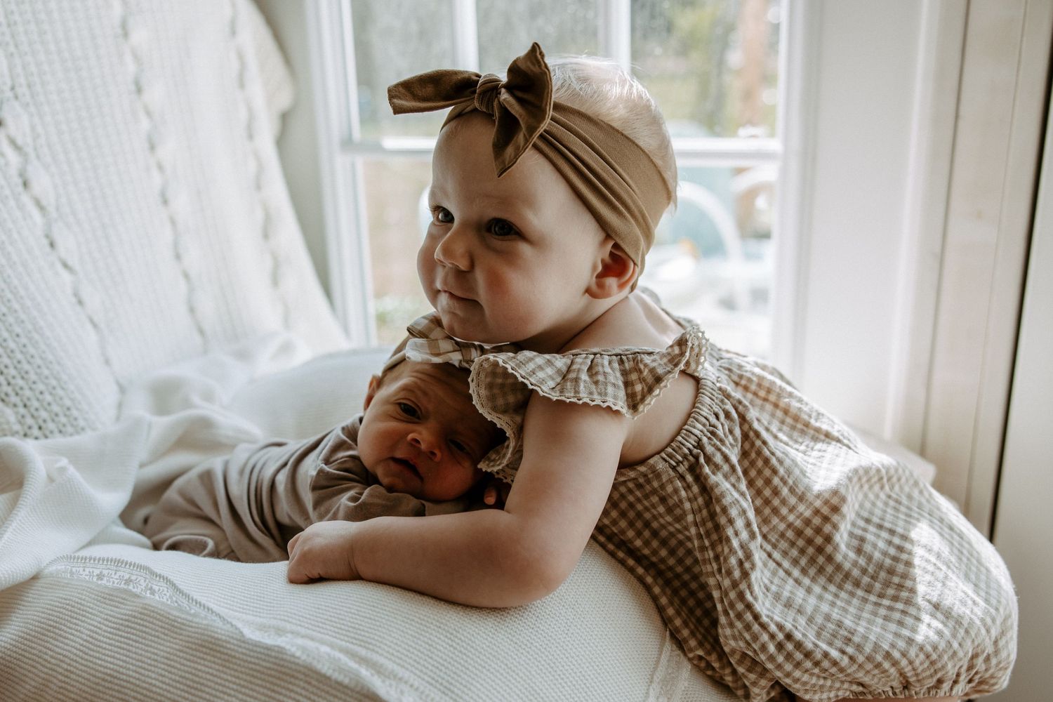 Adorable baby girl with bow headband cuddles a newborn sibling on a white chair by a window.