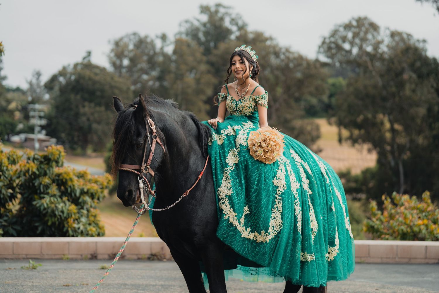 A person in a teal quinceañera dress sits atop a majestic black horse in a garden setting during a photo shoot.