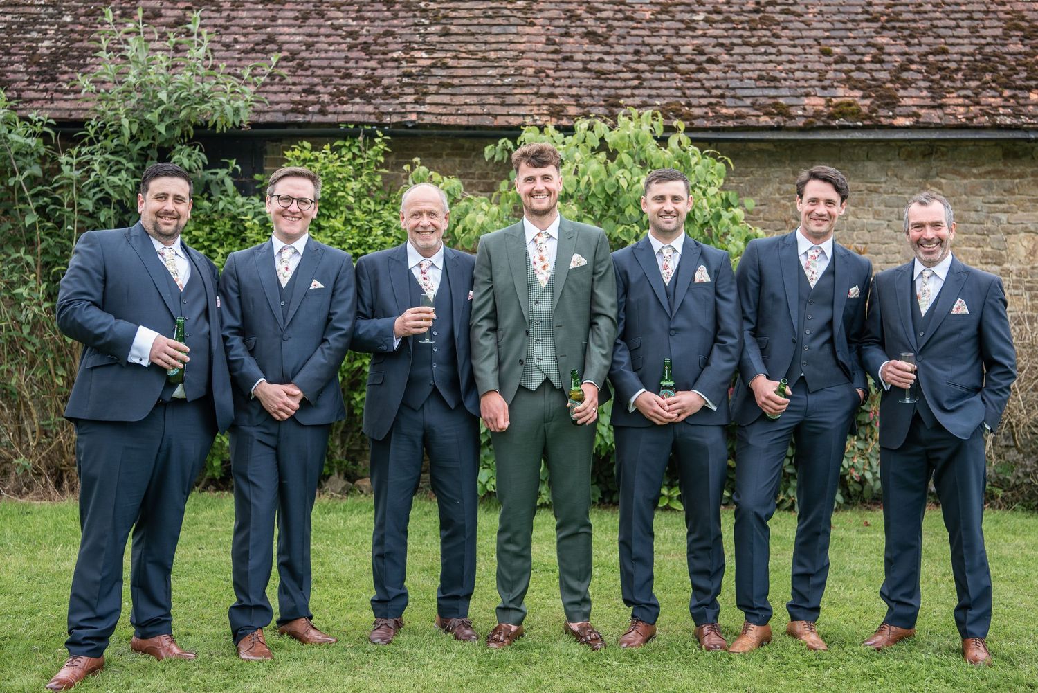 Groom and six friends line up in their suits, many have a beer in hand, they look very relaxed