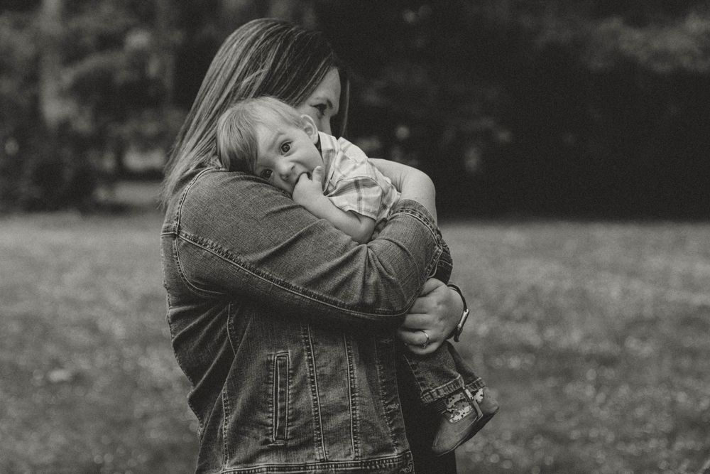 A mother tenderly embraces her baby outdoors in a black and white family portrait photo.