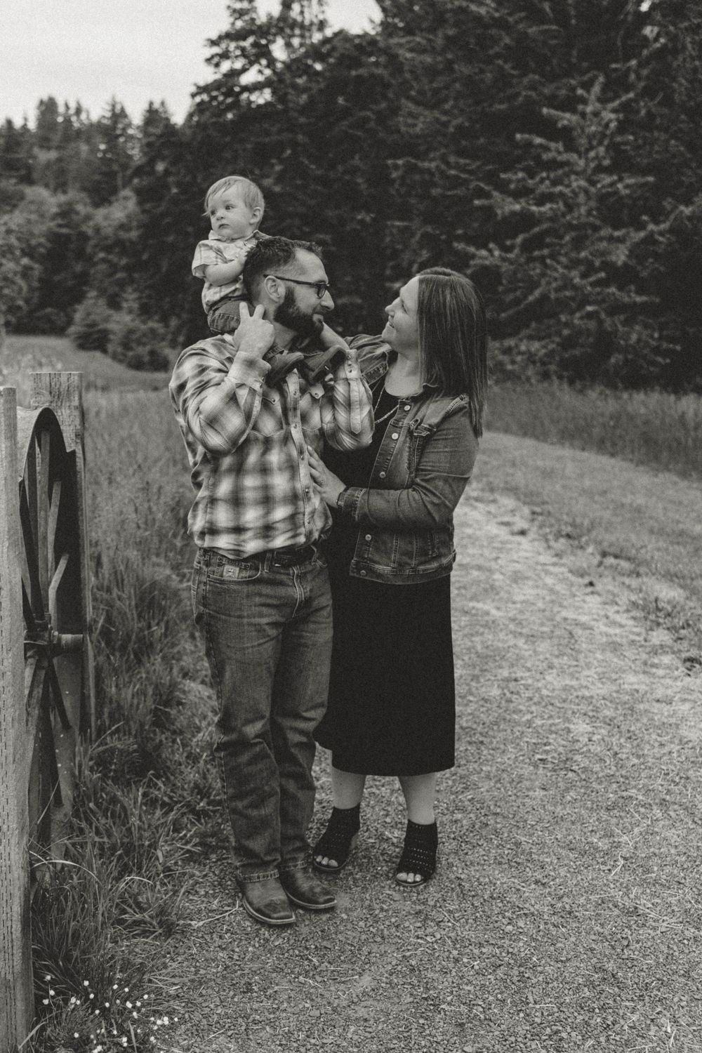 Black and white photo of a couple sharing a tender moment with baby on fathers shoulders outdoors.