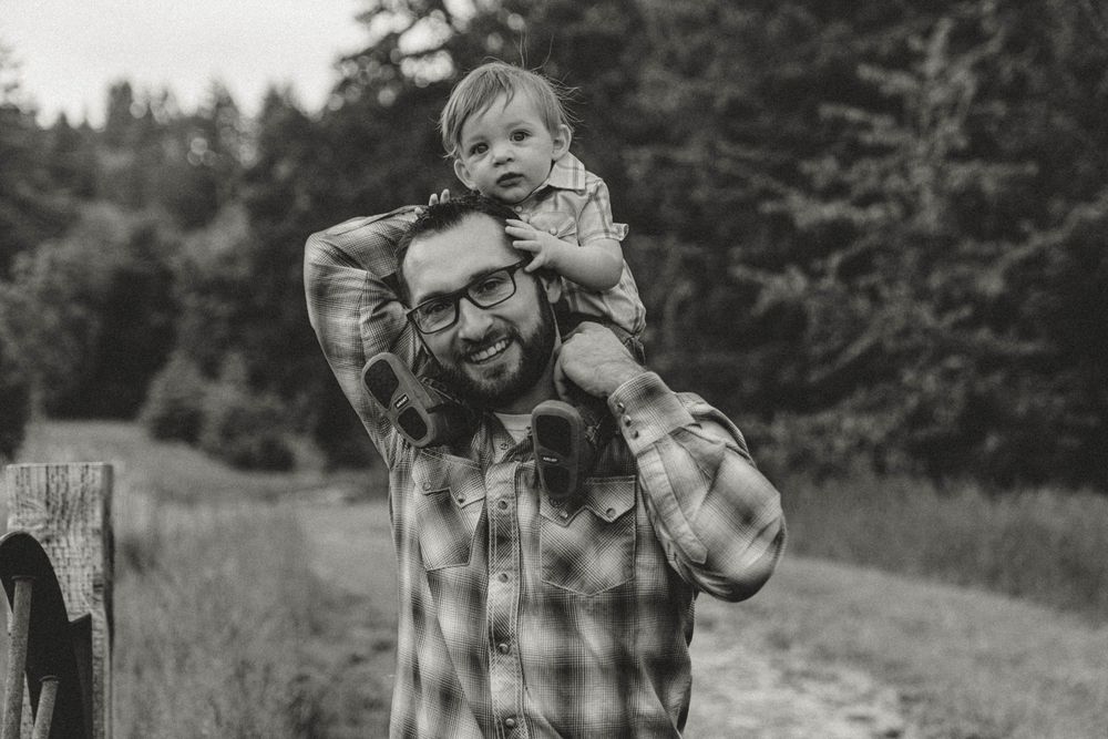 Black and white portrait of a bearded father smiling with his toddler sitting on his shoulders.