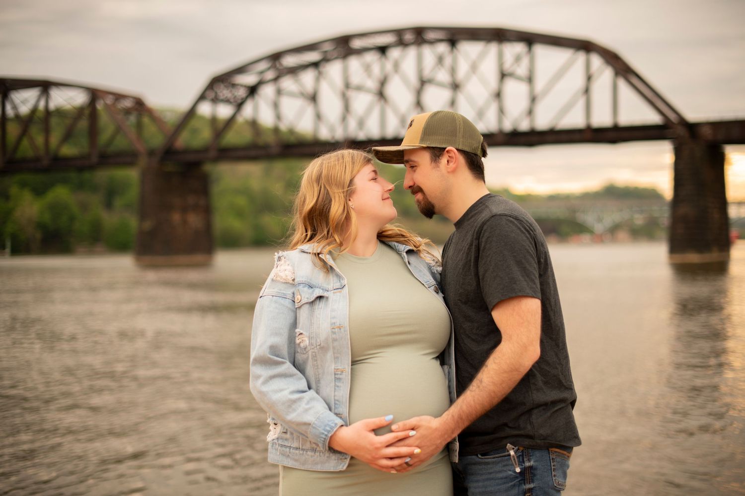 A couple shares a romantic moment by a river during sunset with a rustic bridge in the background.