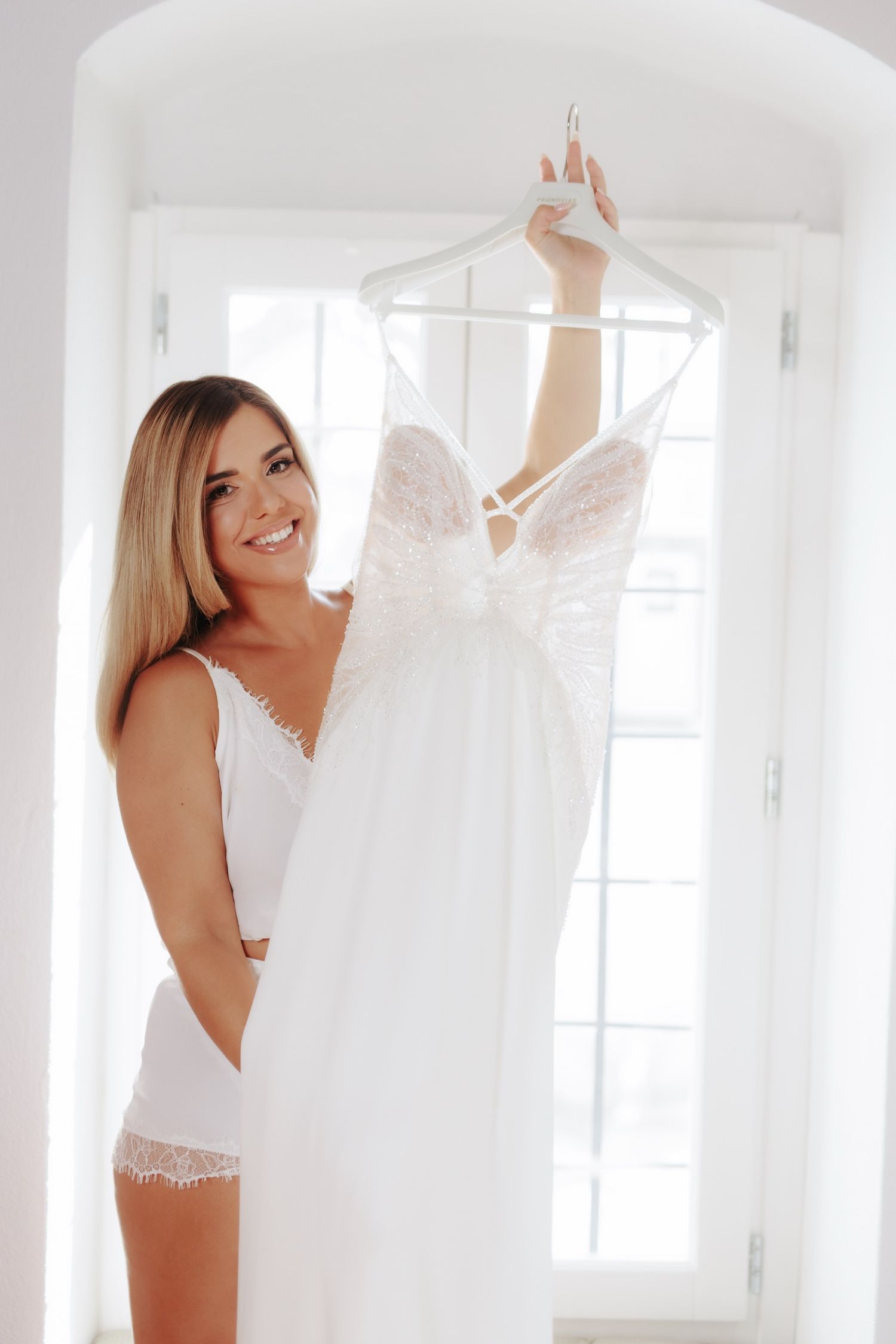 A white wedding dress on a hanger being held up in a bright sunlit room.