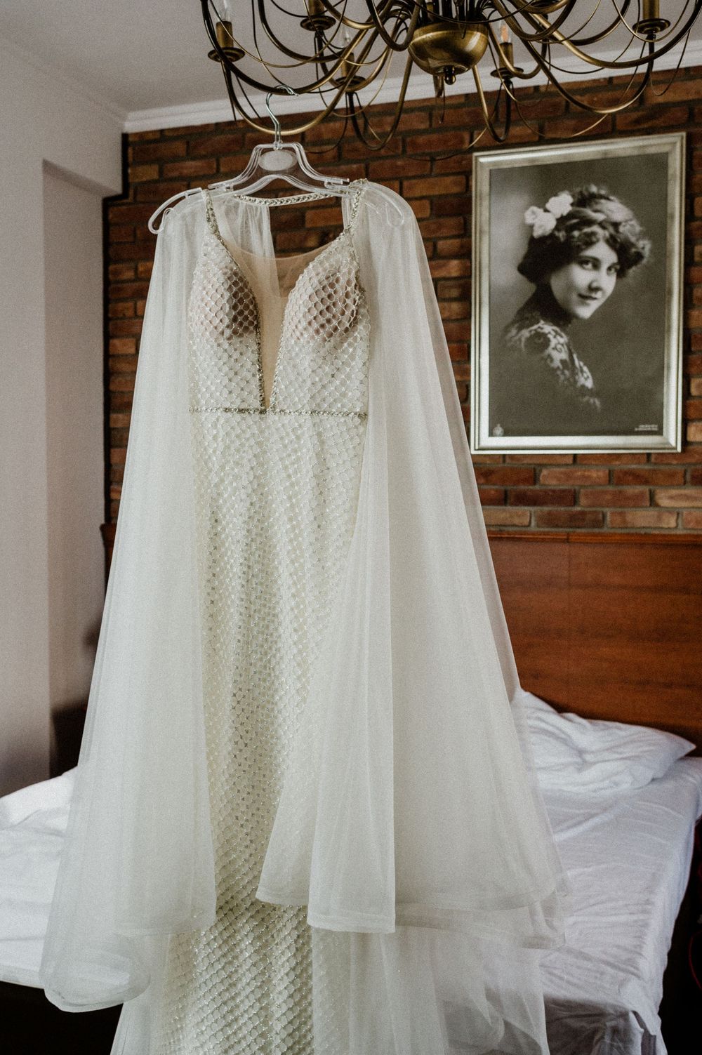 Delicate lace wedding dress with cape hanging against exposed brick wall near vintage portrait.