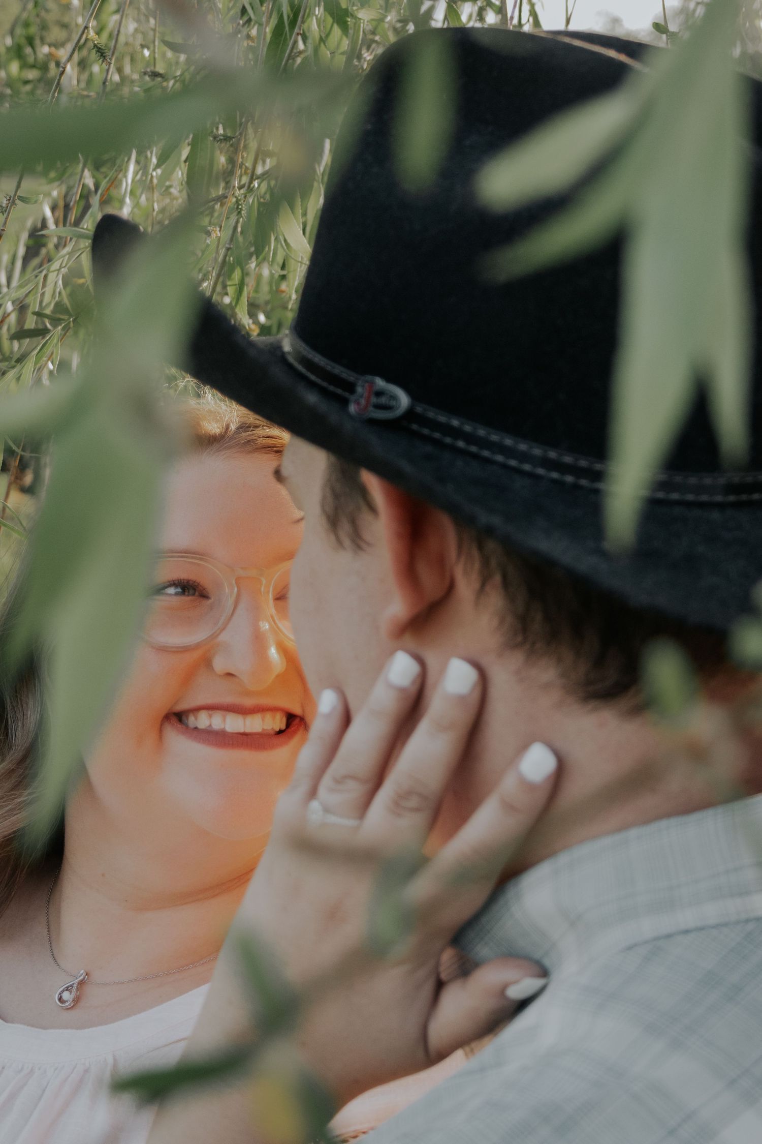 A romantic moment captured through leaves and foliage showing affection between a couple wearing a black cowboy hat.