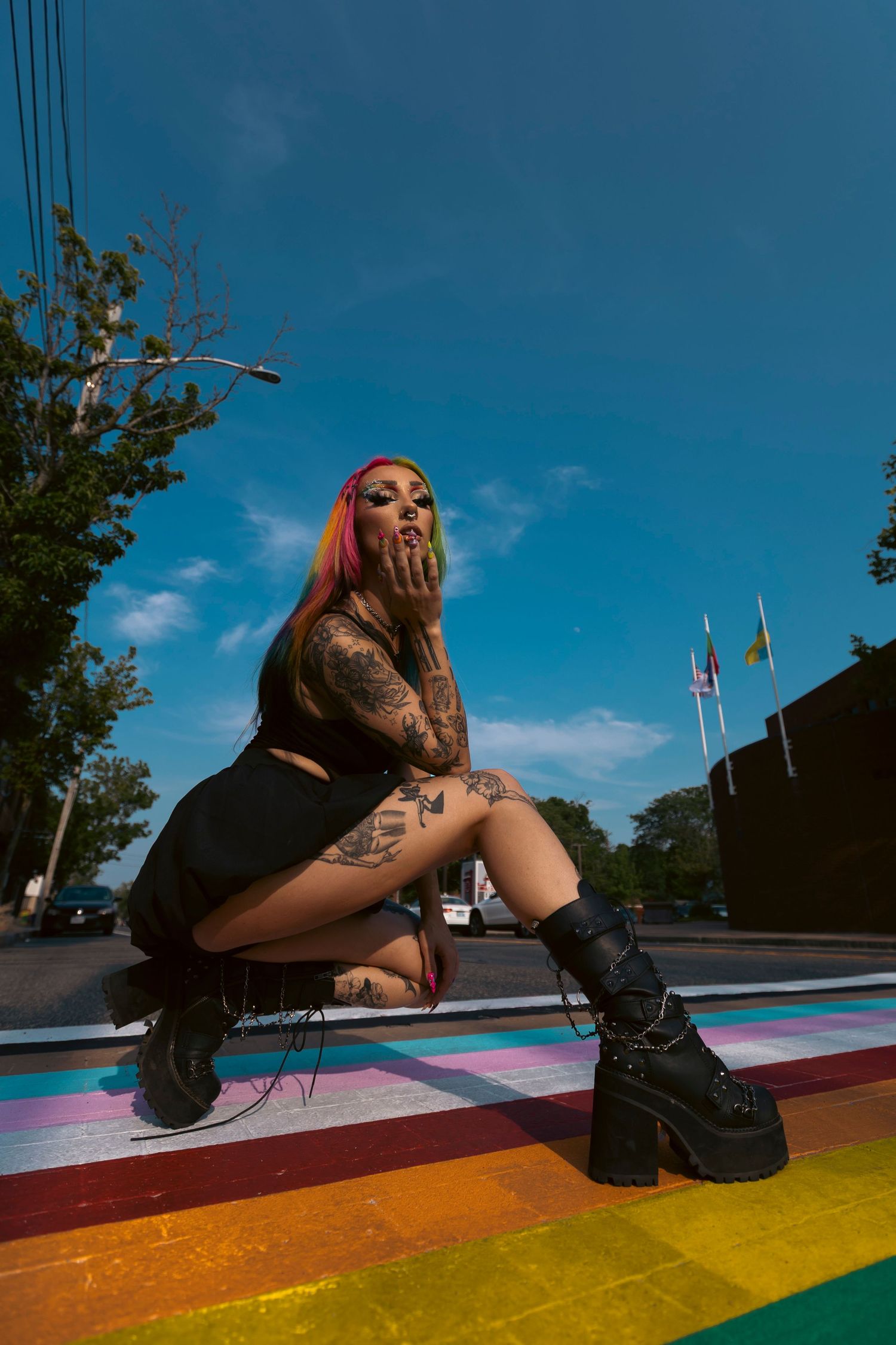 Alternative fashion model in platform boots poses on a rainbow-painted crosswalk against a bright blue sky.