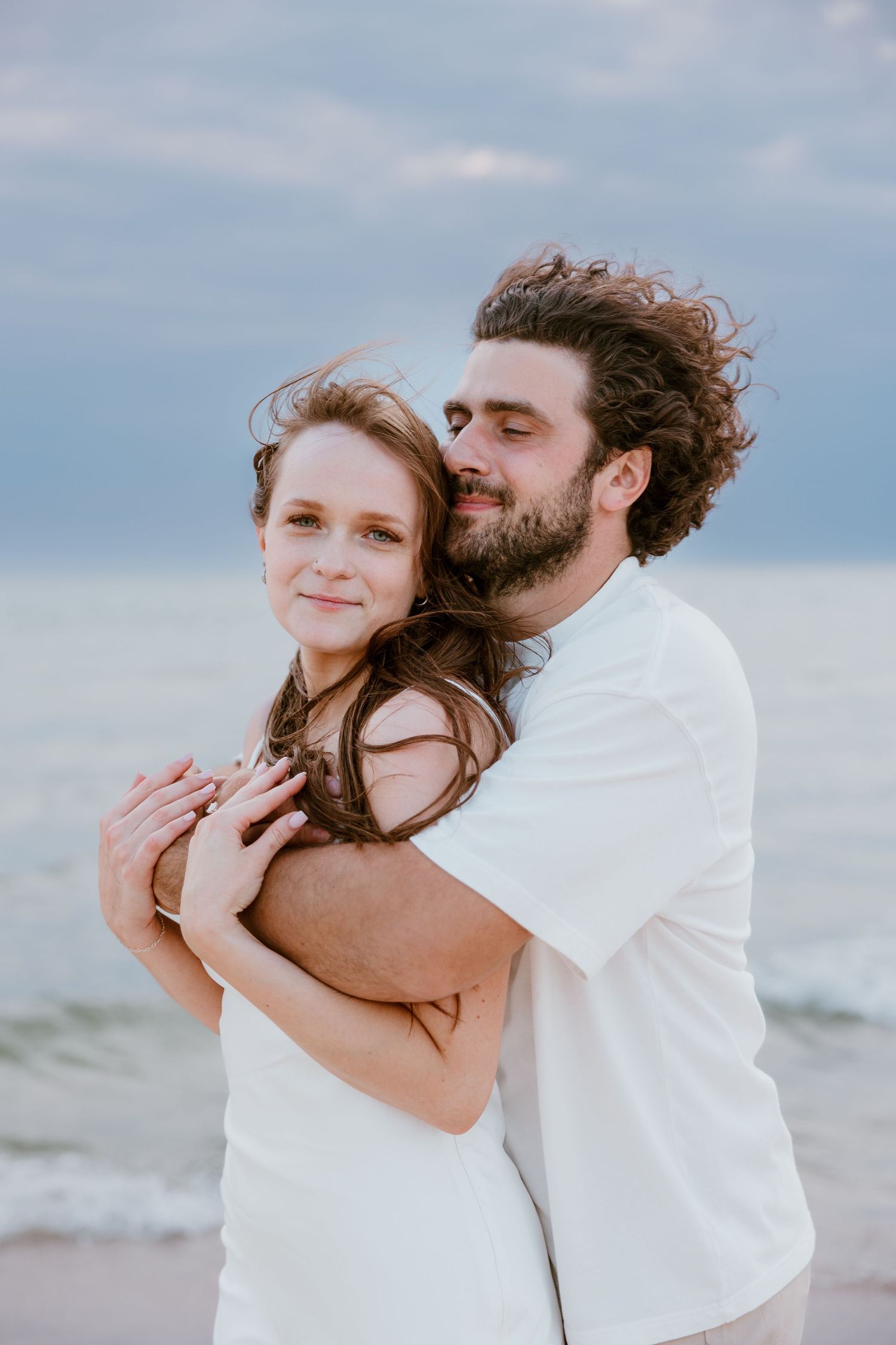 A romantic couple embraces on the beach at dusk with soft lighting and ocean view in the background. Michigan Wedding Photographer