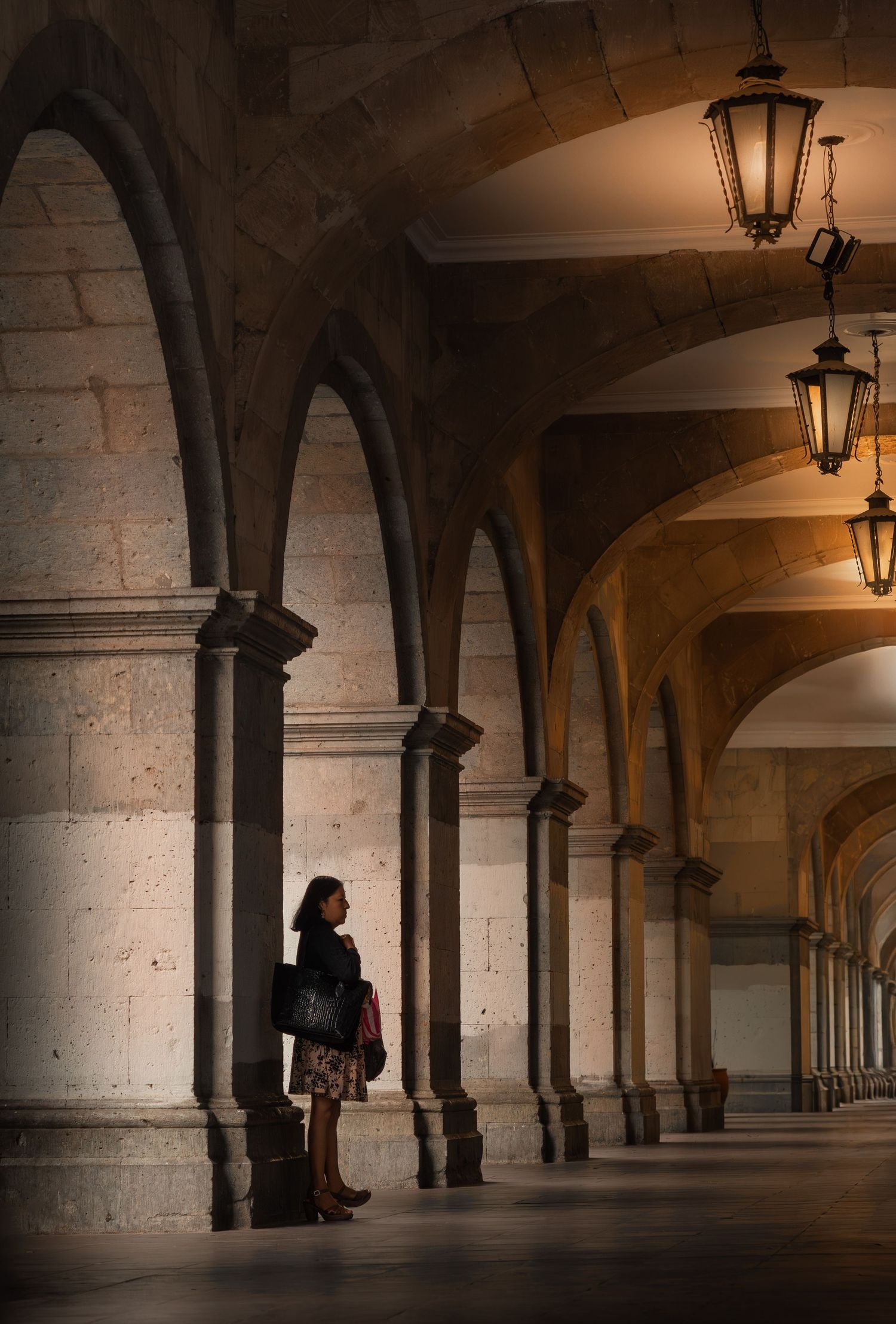 A dark atmospheric colonnade with stone arches and hanging lanterns creates a moody architectural perspective in Oaxaca, Mexico.