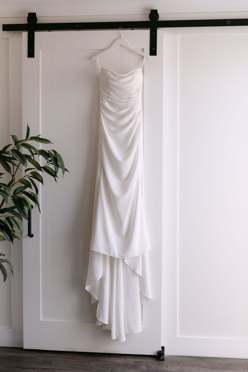 Four white wedding dresses displayed on hangers against a white wall with minimal green plant decor.