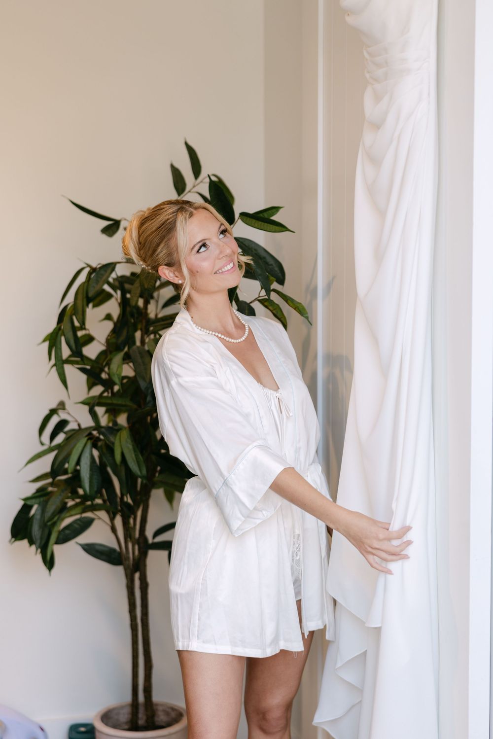 Woman in white silk robe standing by window curtains with indoor plant in elegant bright interior.