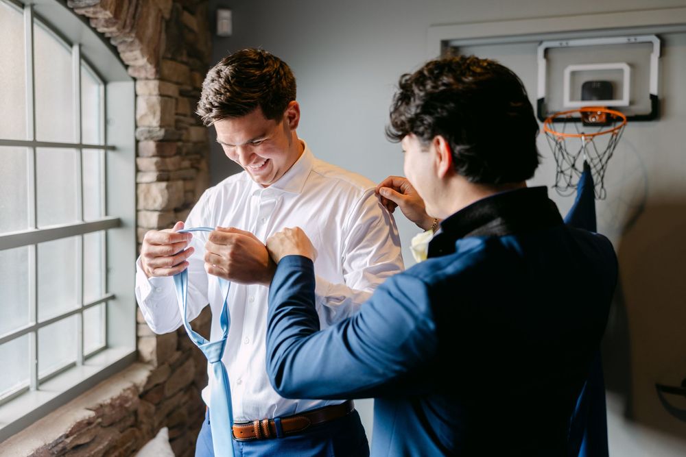 Two people sharing a joyful moment while getting dressed in formal attire near a window.