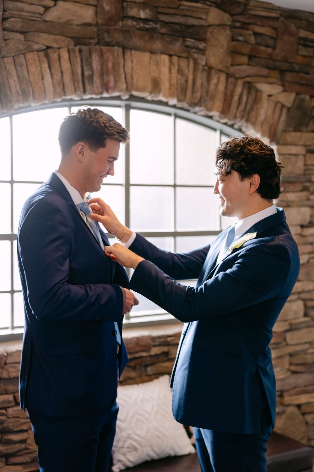 Two groomsmen in dark suits adjusting each other's ties in a stone-walled room with windows.