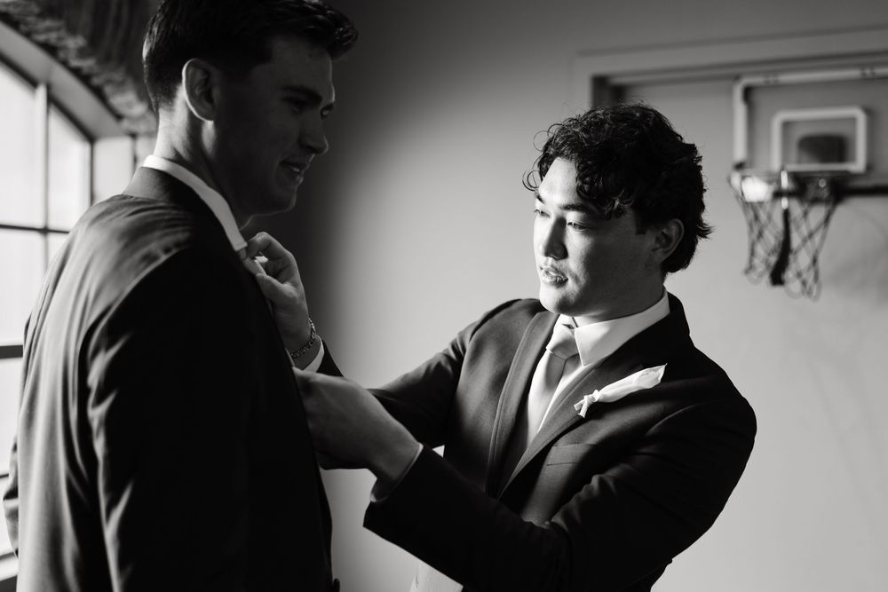 Black and white photo of two people adjusting formal wear in a room with natural lighting.