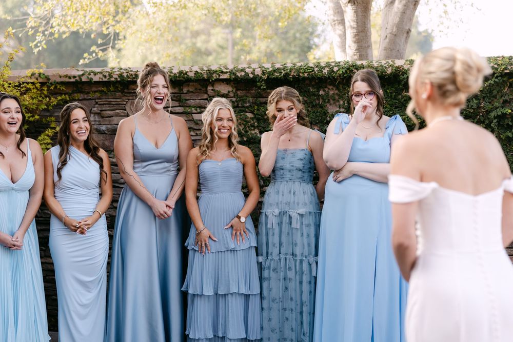 Bridesmaids wearing light blue dresses stand together in a row outdoors.