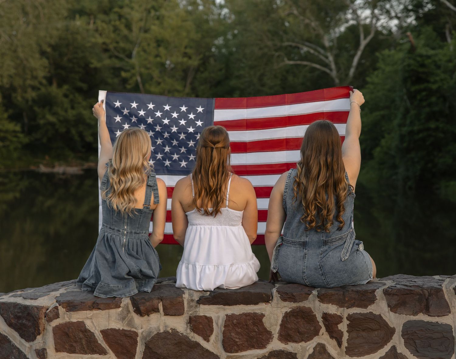 Three girls sitting on a stone wall holding an American flag with their backs to the camera near a river.