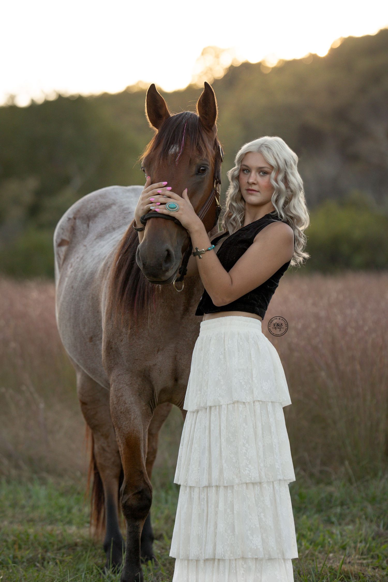 A brown horse and someone in a white tiered dress stand together in a grassy field at sunset.