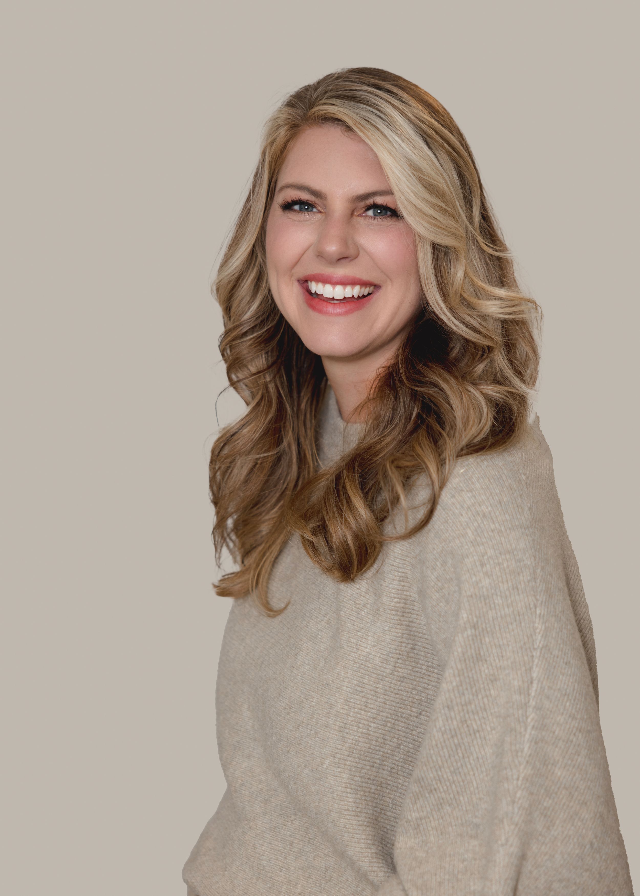 Woman in beige sweater smiling against neutral background.