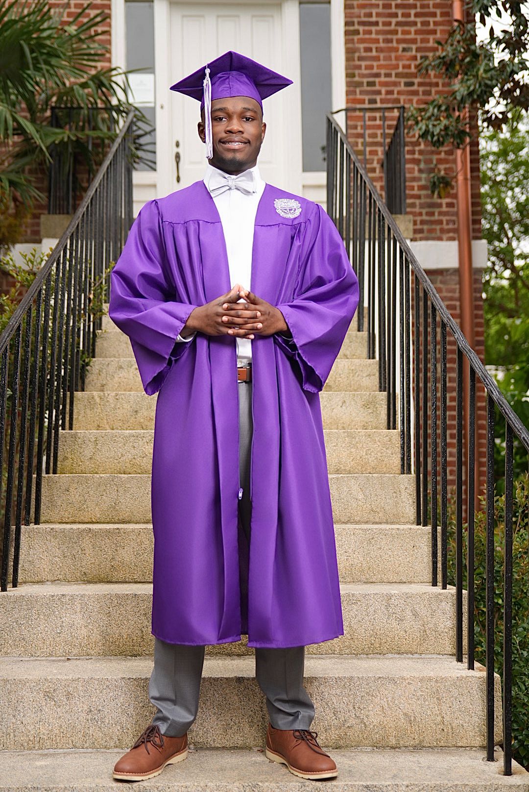 Graduate in a purple cap and gown with a bow tie and brown shoes poses with hands clasped on brick steps with black iron railings.