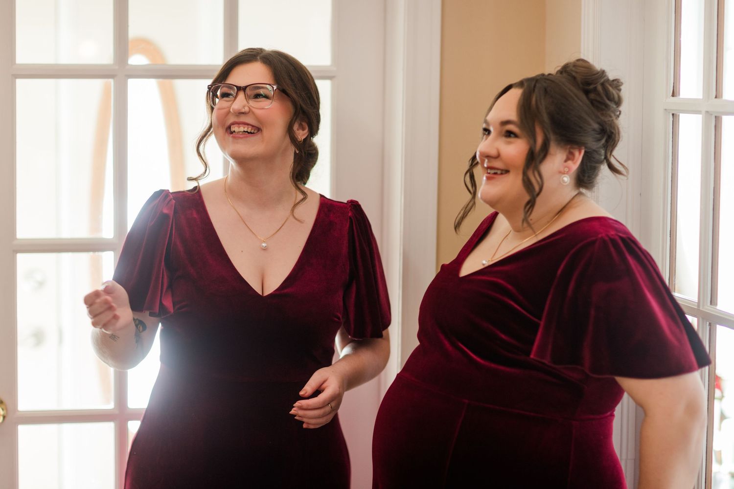 A group wearing burgundy velvet dresses laughs and dances together in a bright indoor space.