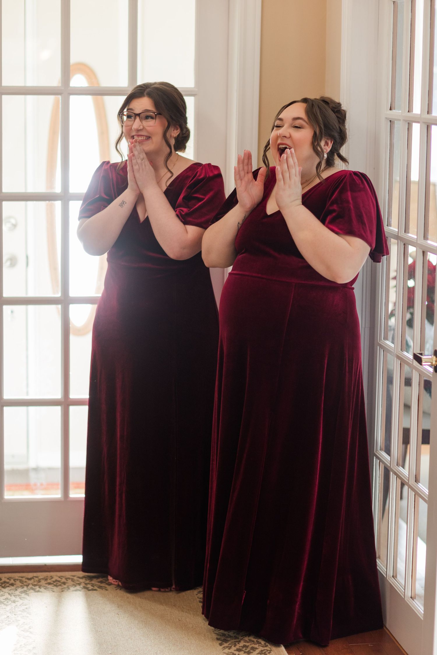 A group of bridesmaids in burgundy velvet dresses pose playfully in a sunlit hallway.