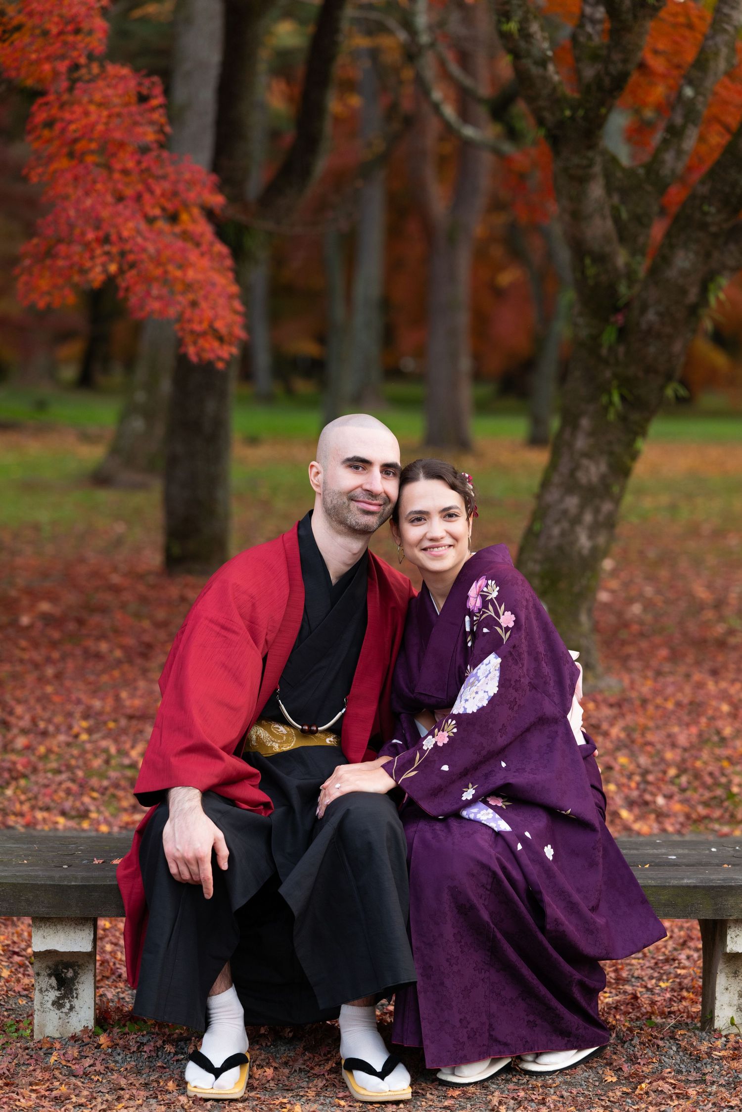 Couple in traditional Japanese kimono sitting on bench surrounded by vibrant red autumn foliage in a Japanese garden.