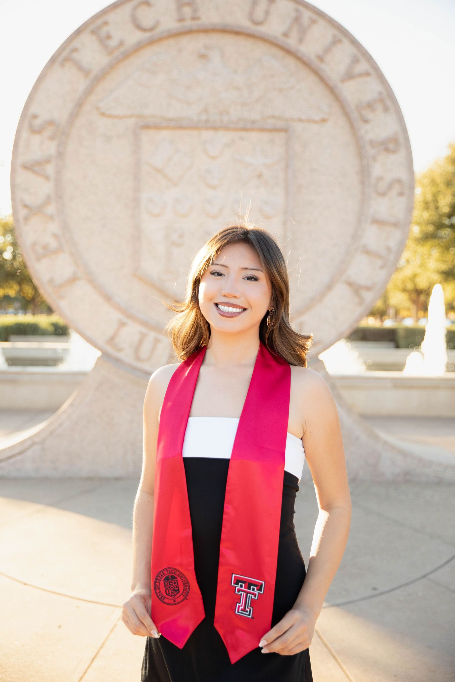 Graduate poses in black dress and red stole in front of university seal during golden hour sunset.