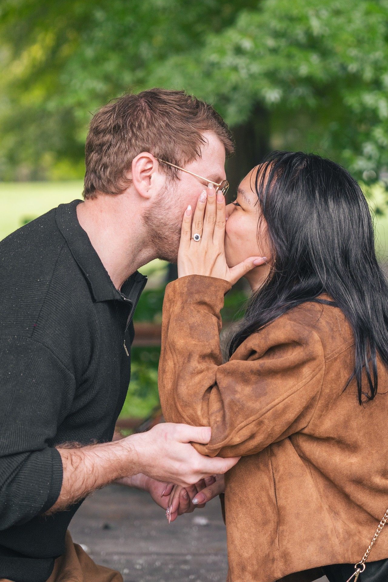 A couple shares an intimate moment outdoors as she whispers something in his ear while wearing a brown jacket.