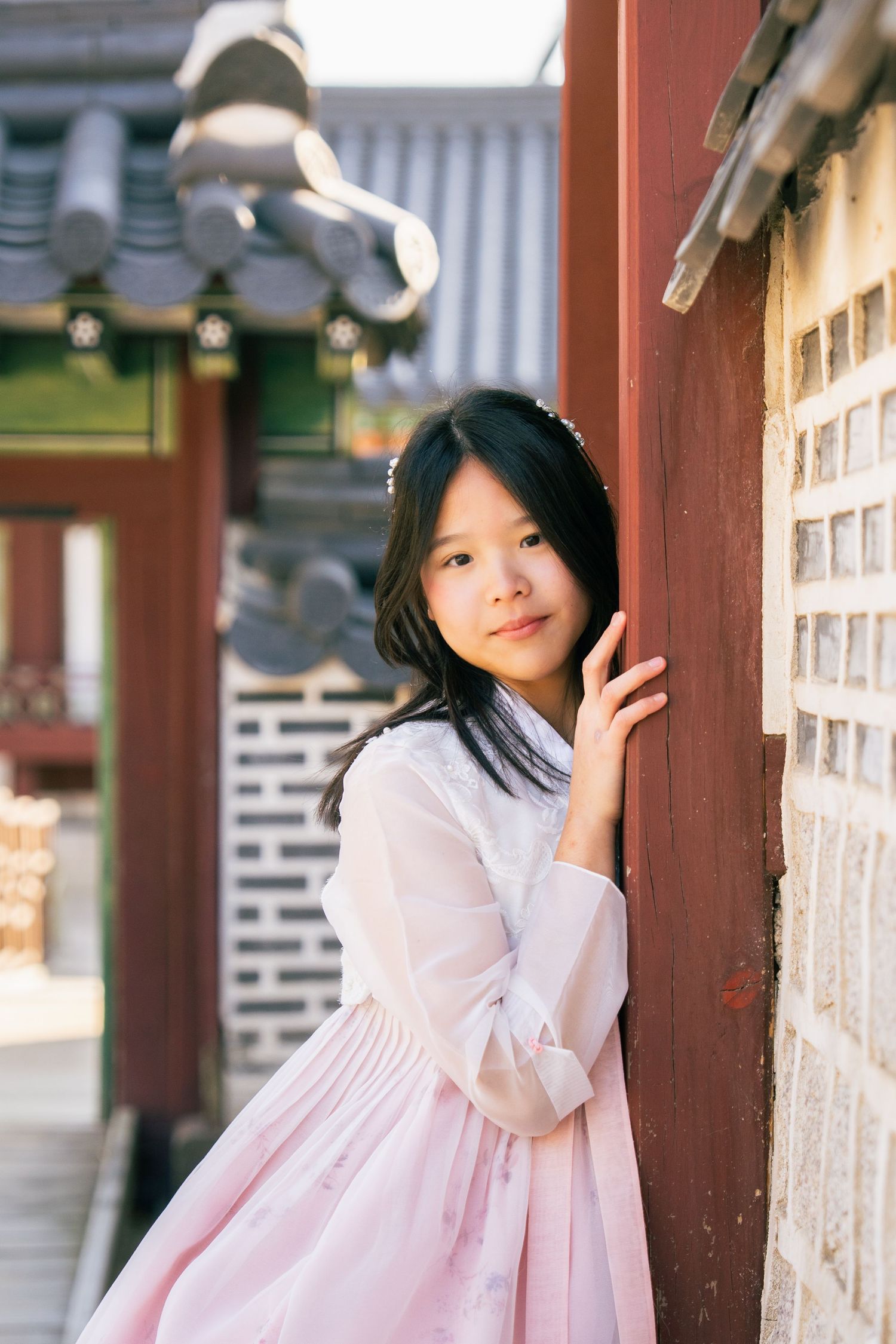 A person in a pink hanbok dress poses against a traditional Korean architectural wall with lanterns in the background.