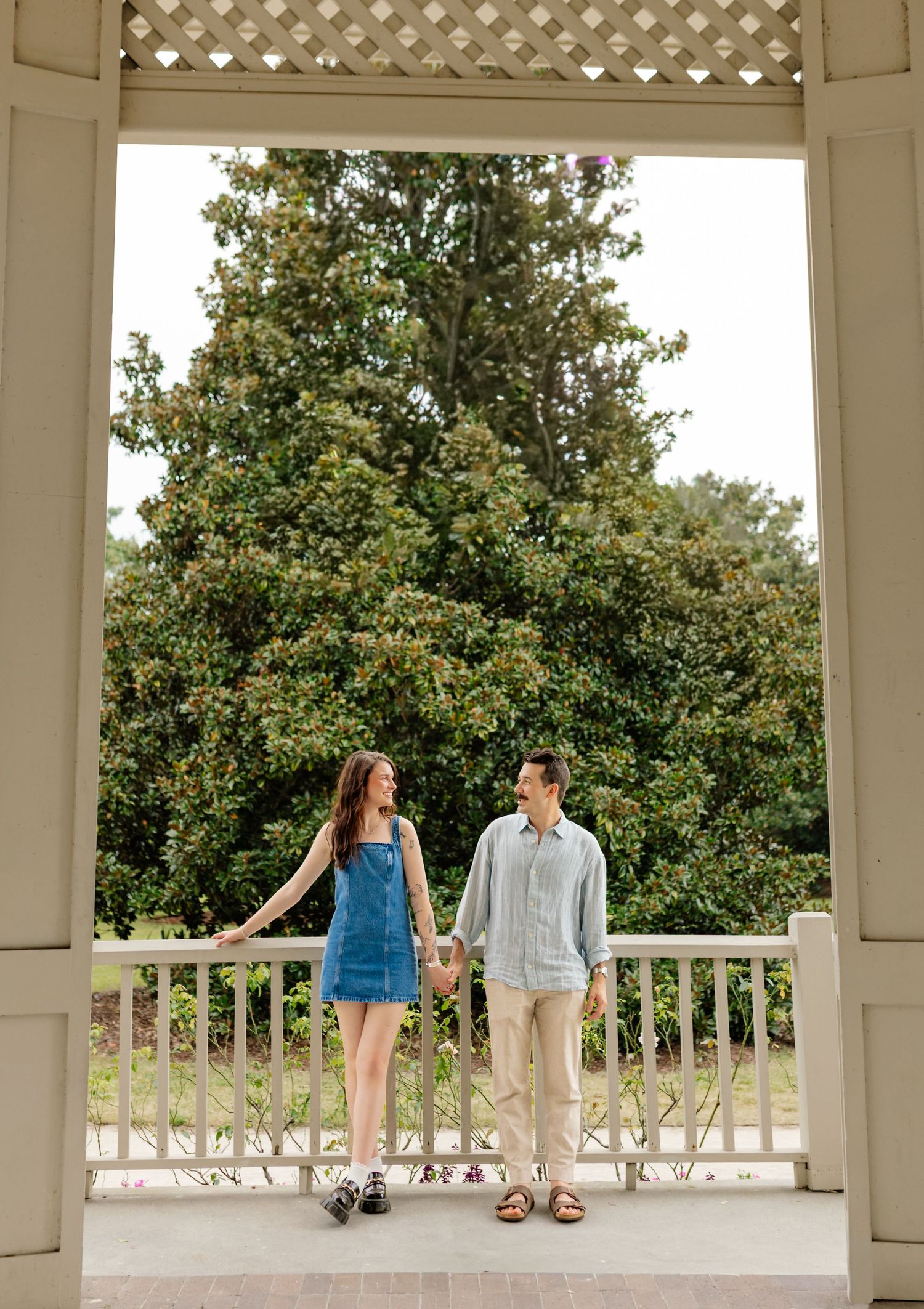 Couple sitting and walking along white gazebo railing in casual summer outfits.