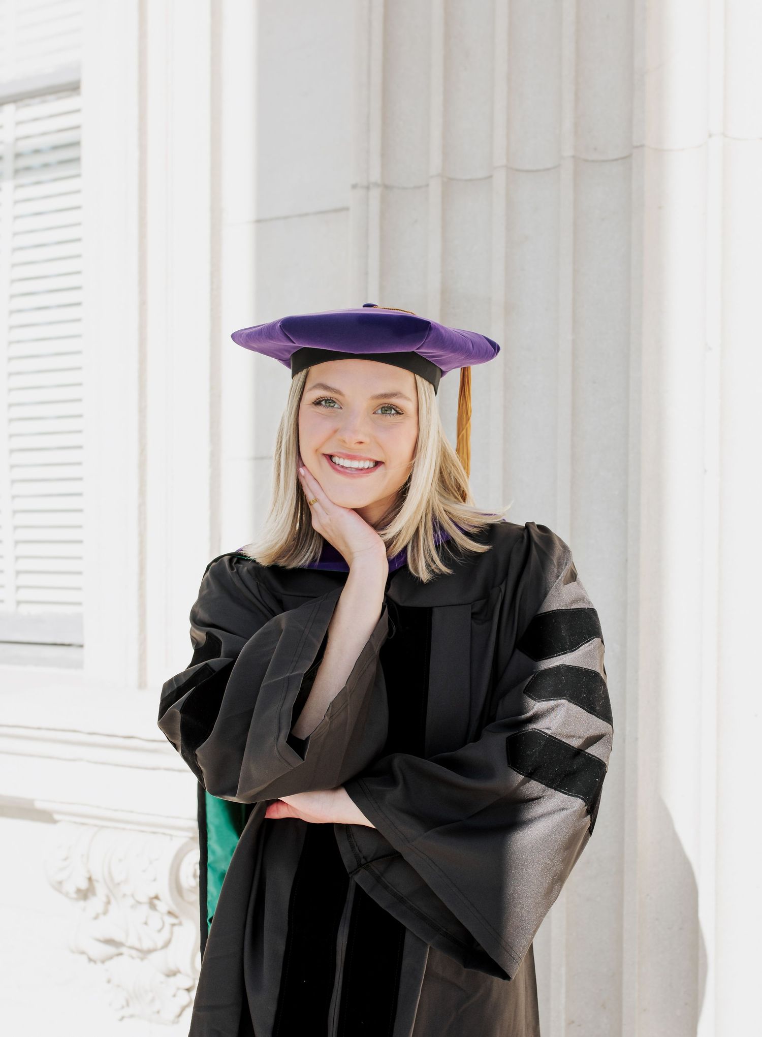 Graduate in cap and gown poses for portraits against white columns.