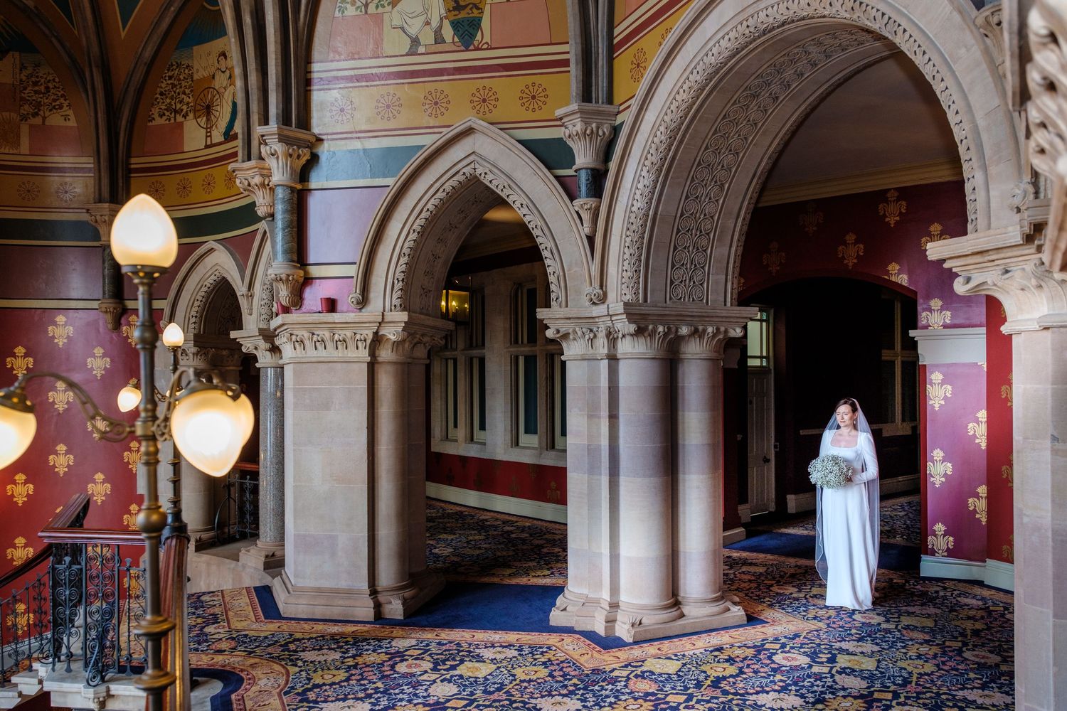 Gothic stone archways and ornate Victorian interior with patterned carpet and decorative walls in historic mansion hallway.