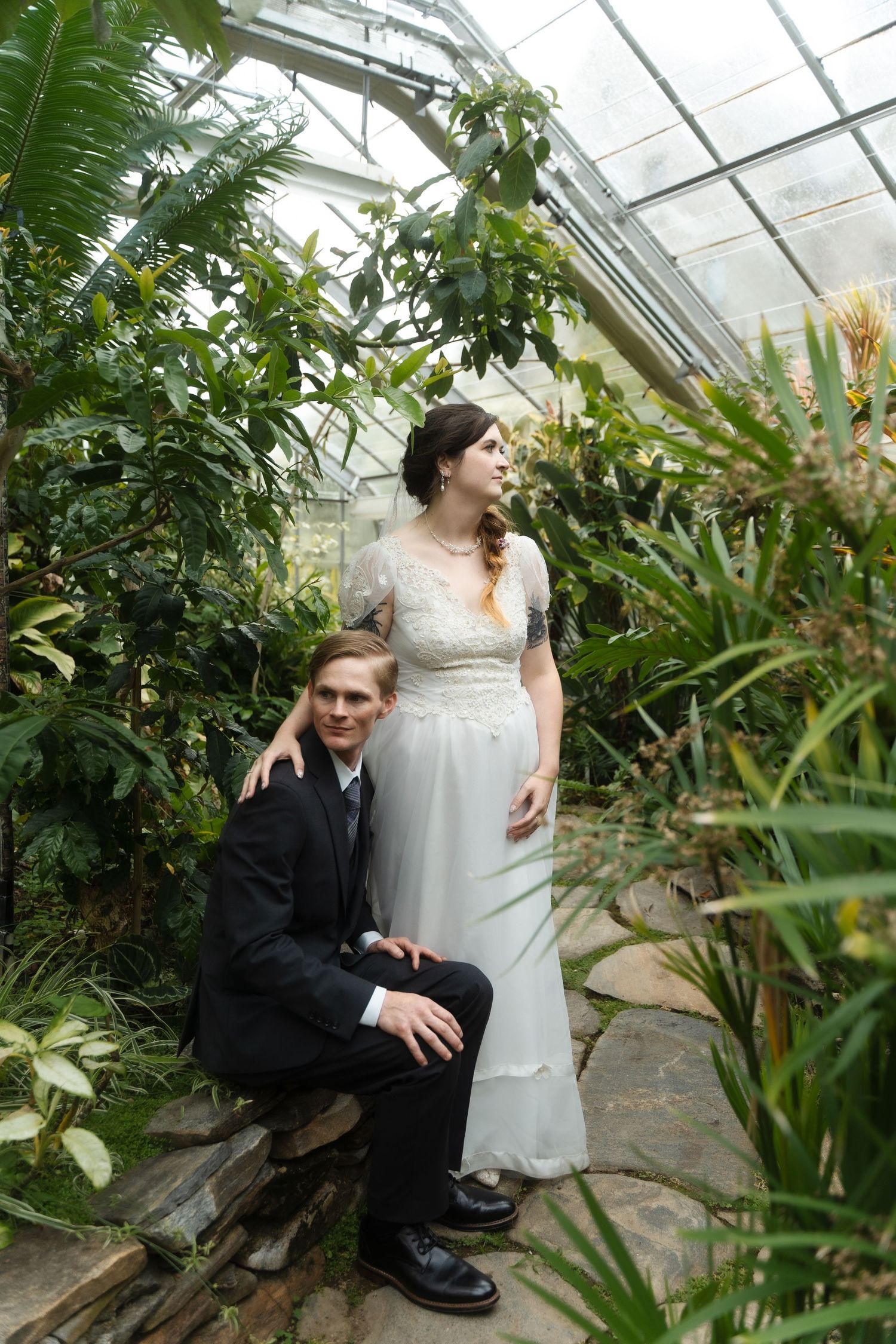 A couple poses for wedding photos in a lush greenhouse conservatory filled with tropical plants and natural light.