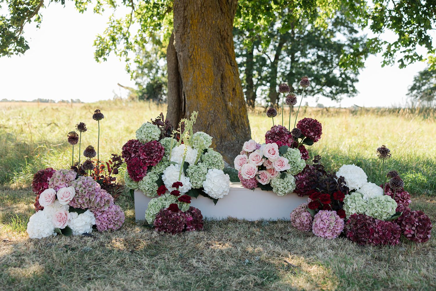 A rustic outdoor wedding arch decorated with pink and burgundy flowers stands against a tree in a countryside setting.
