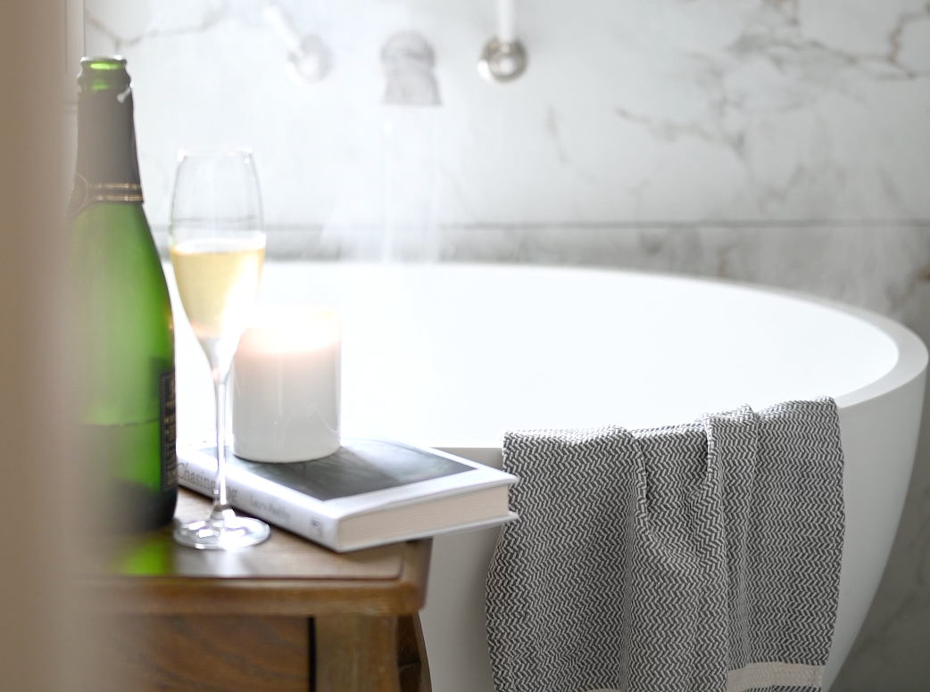 A modern bathroom interior with marble walls, grey towels, and a sleek white bathtub near a window, with a glass of wine and steam from the