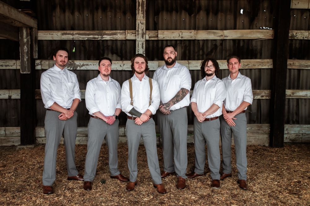Groomsmen in matching grey pants and white shirts pose together in a rustic barn setting.